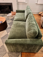 A green velvet sofa is positioned against a wall. A round wooden coffee table sits nearby, with a patterned rug beneath. Cozy pillows add warmth to the space.