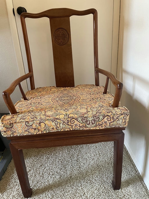 A wooden chair with a carved backrest and intricately patterned fabric cushion sits on beige carpeting, against a light-colored wall.