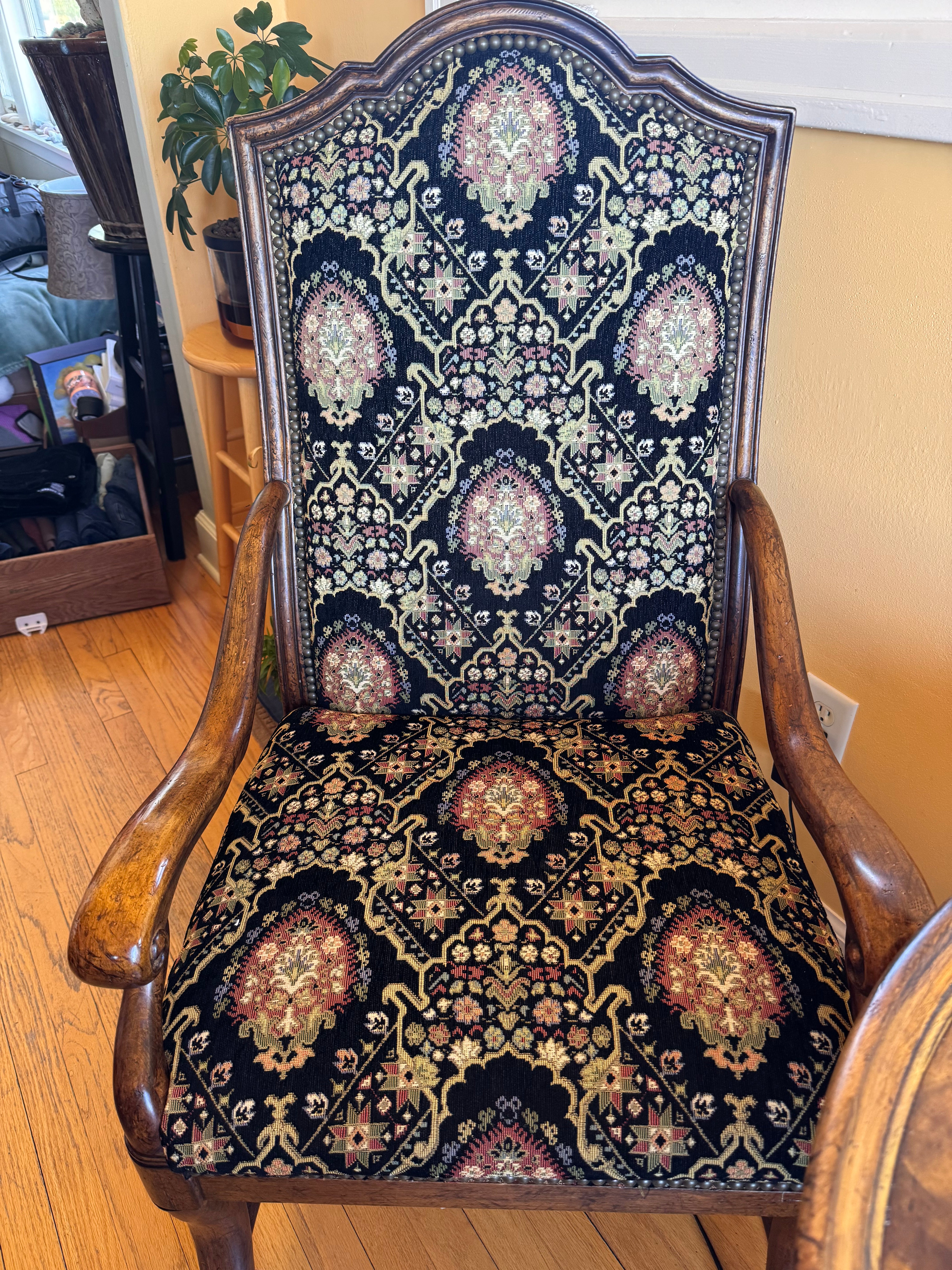 A vintage wooden chair with intricate floral-patterned upholstery in black, red, and gold. The chair features curved armrests and detailed craftsmanship.