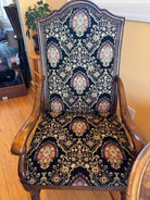 A vintage wooden chair with intricate floral-patterned upholstery in black, red, and gold. The chair features curved armrests and detailed craftsmanship.