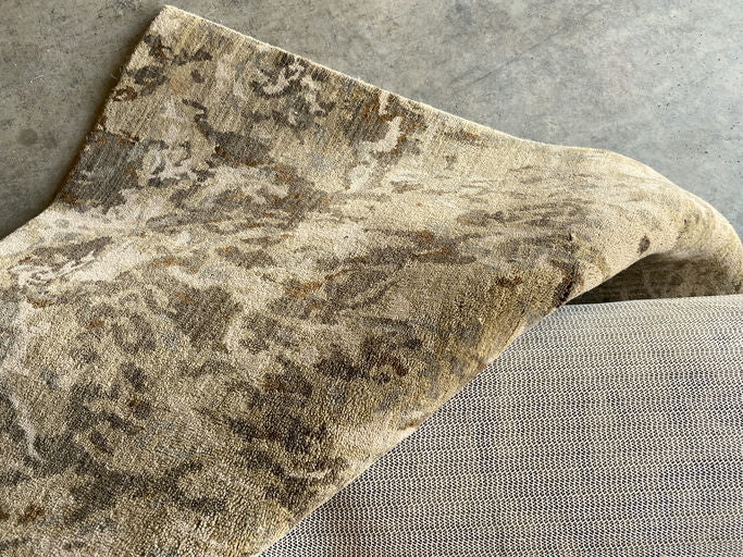 A patterned, light-colored rug is partially draped over a textured, neutral fabric surface, showcasing intricate designs against a concrete floor.