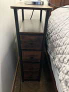 A wooden nightstand with four small drawers and a flat surface on top, housing a lamp. Positioned next to a bed. Wall and carpet visible.