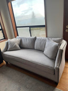 A light gray sofa with two decorative pillows, positioned near large windows that provide a view of the outdoors. The floor is wooden.