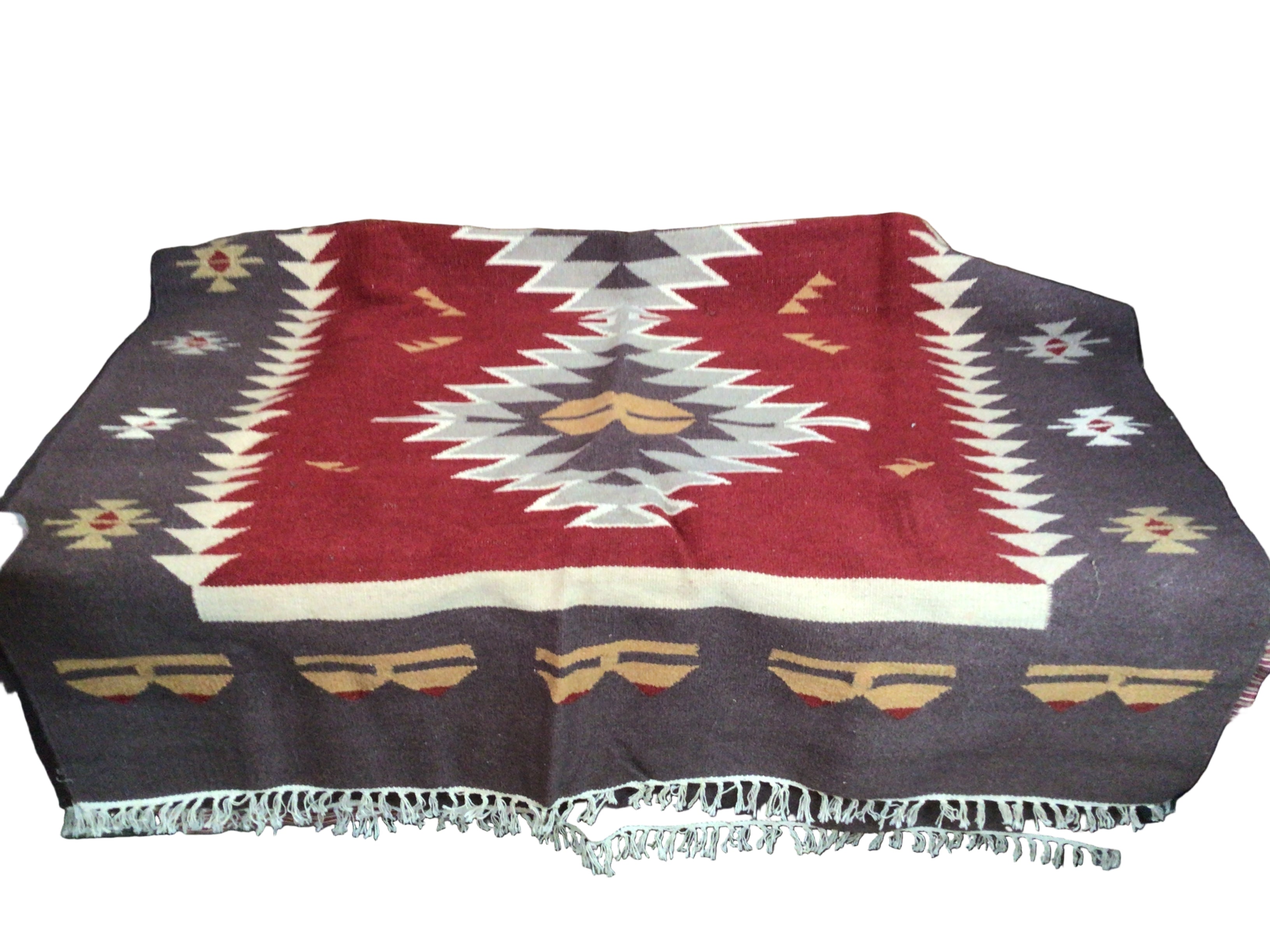 A patterned blanket featuring geometric designs in red, cream, and brown, laid on a surface, with fringed edges and a rustic wooden background.