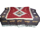 A patterned blanket featuring geometric designs in red, cream, and brown, laid on a surface, with fringed edges and a rustic wooden background.