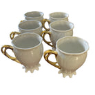 A row of six elegant white ceramic cups with gold handles, displayed on a wooden table. The cups have decorative bases and are empty.