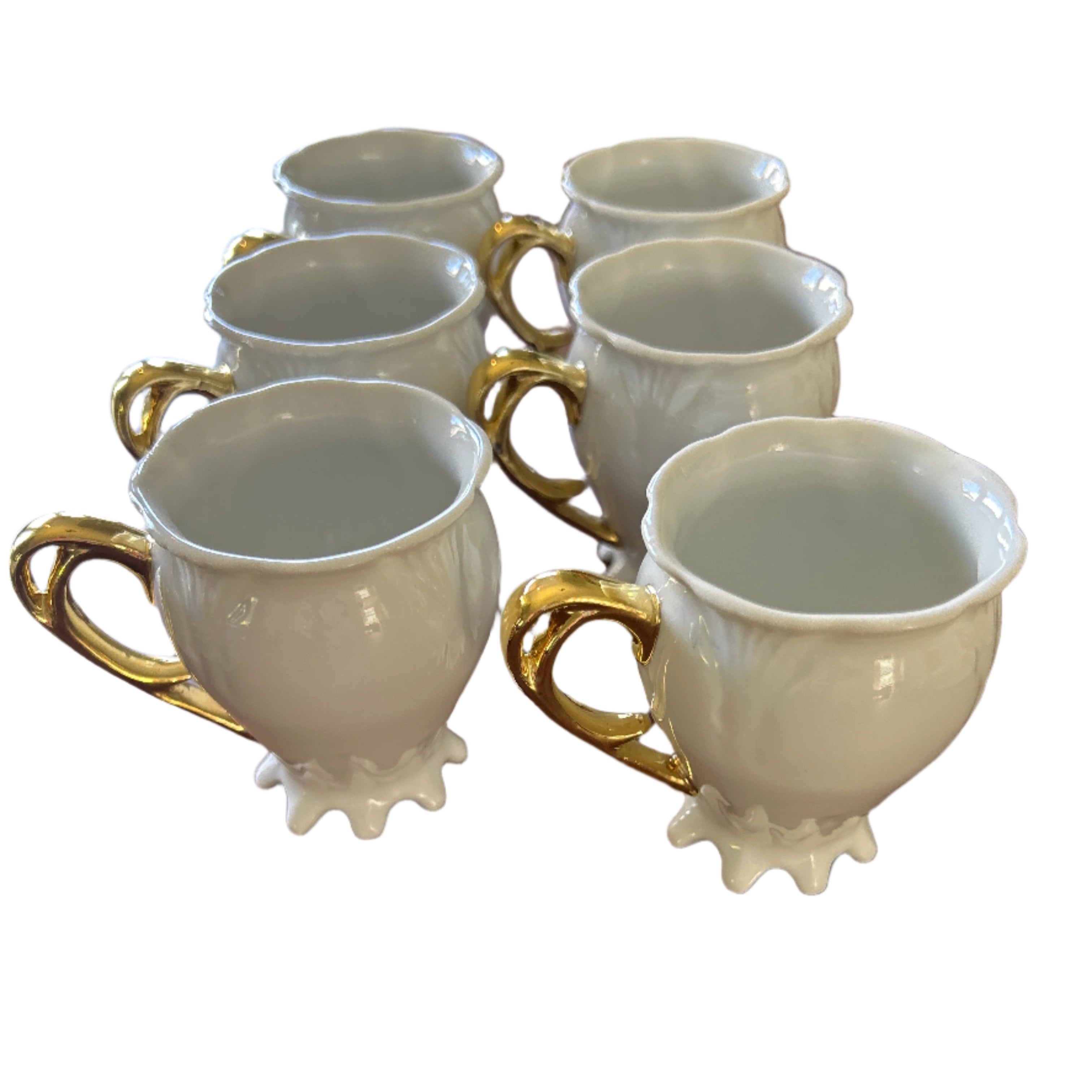 A row of six elegant white ceramic cups with gold handles, displayed on a wooden table. The cups have decorative bases and are empty.