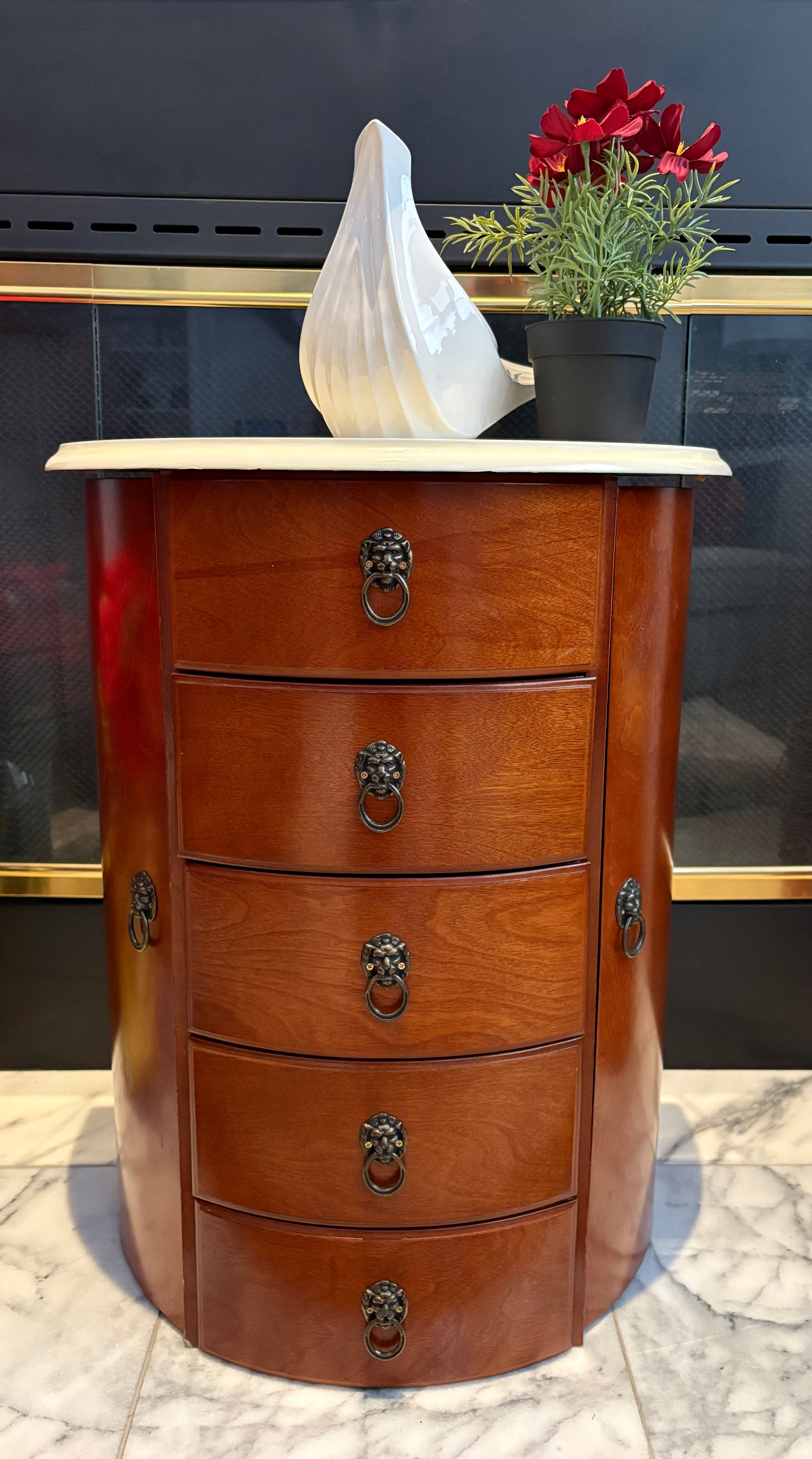 A round wooden cabinet with five drawers, featuring lion's head handles. On top, a white decorative object and a potted plant with red flowers.