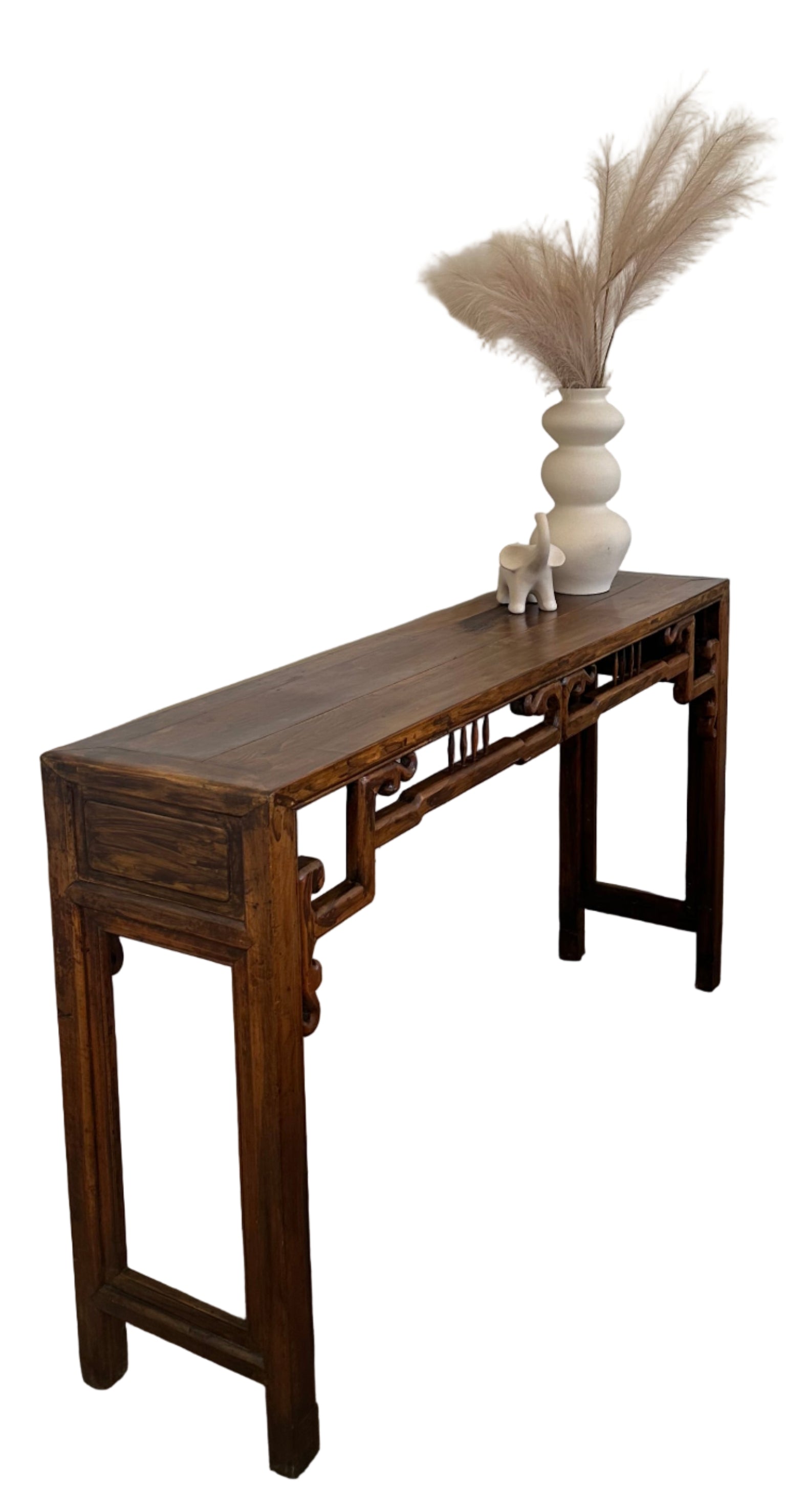A wooden console table with intricate details stands against a white wall, adorned with a white vase and decorative pampas grass. A gray sofa is nearby.