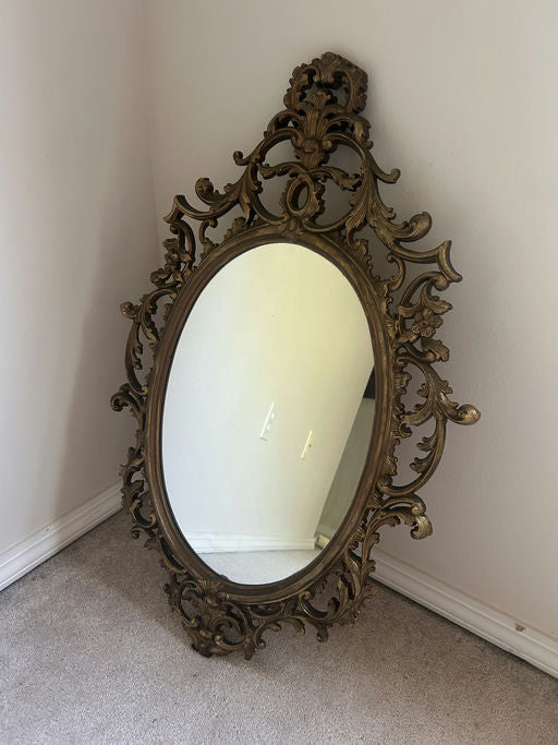 An ornate, vintage mirror with an elegant gold frame rests against a wall on soft carpet, reflecting the room's subtle details.
