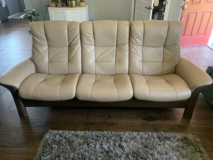 A light beige leather sofa with three seats and plush backing, sitting on a wooden floor with a textured rug in front, and a decorated shelf in the background.