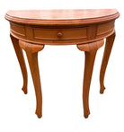 A curved, wooden console table with one drawer and elegant, tapered legs, placed on concrete near a gray corrugated backdrop.