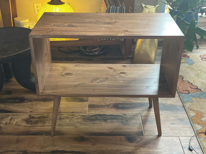 A wooden side table with an open storage space and tapered legs, situated in a cozy room with a plant and decorative lamp in the background.