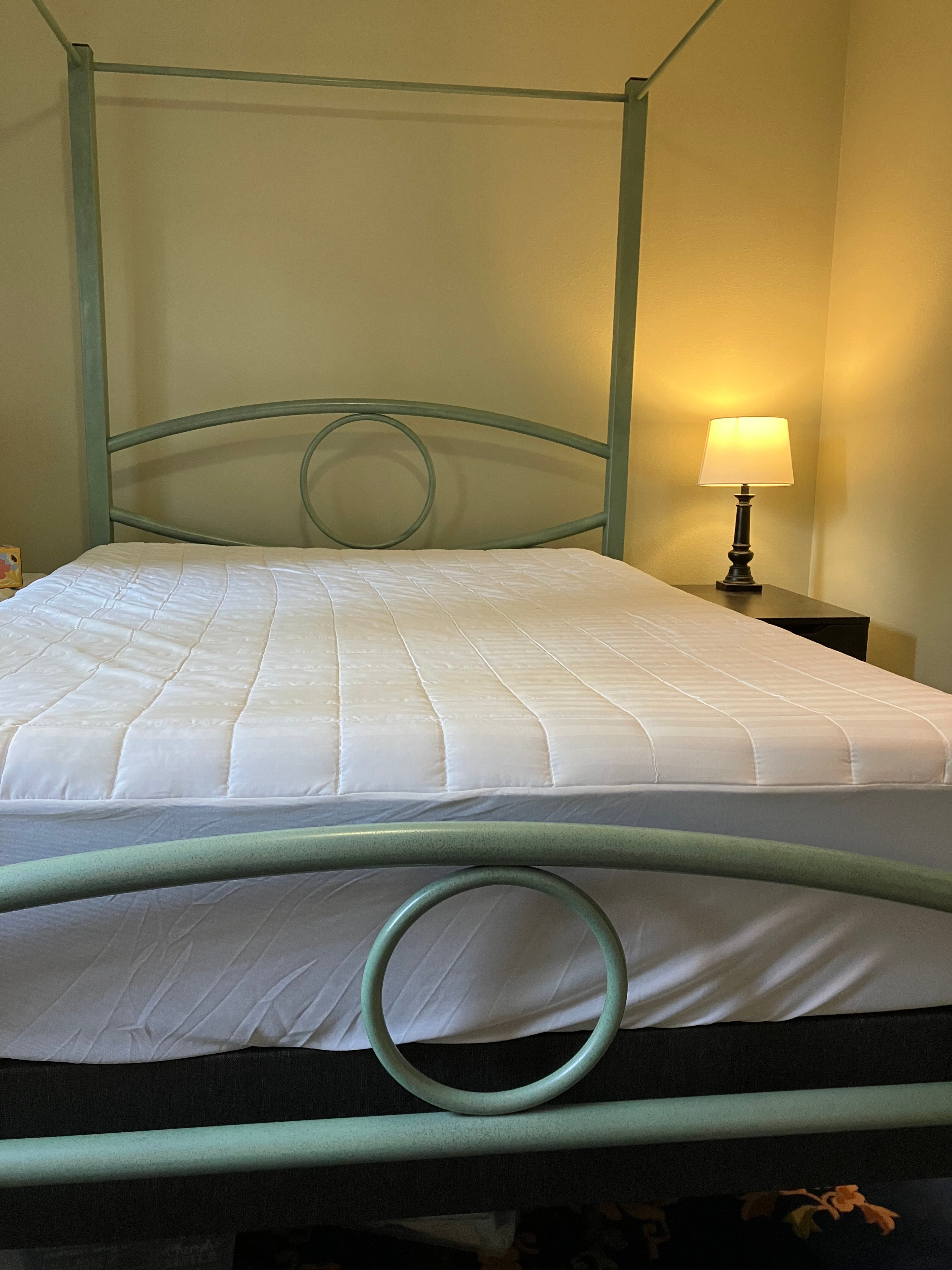 A green metal bed frame with a canopy holds a white mattress. A bedside lamp casts a warm glow in the softly lit room with neutral walls.