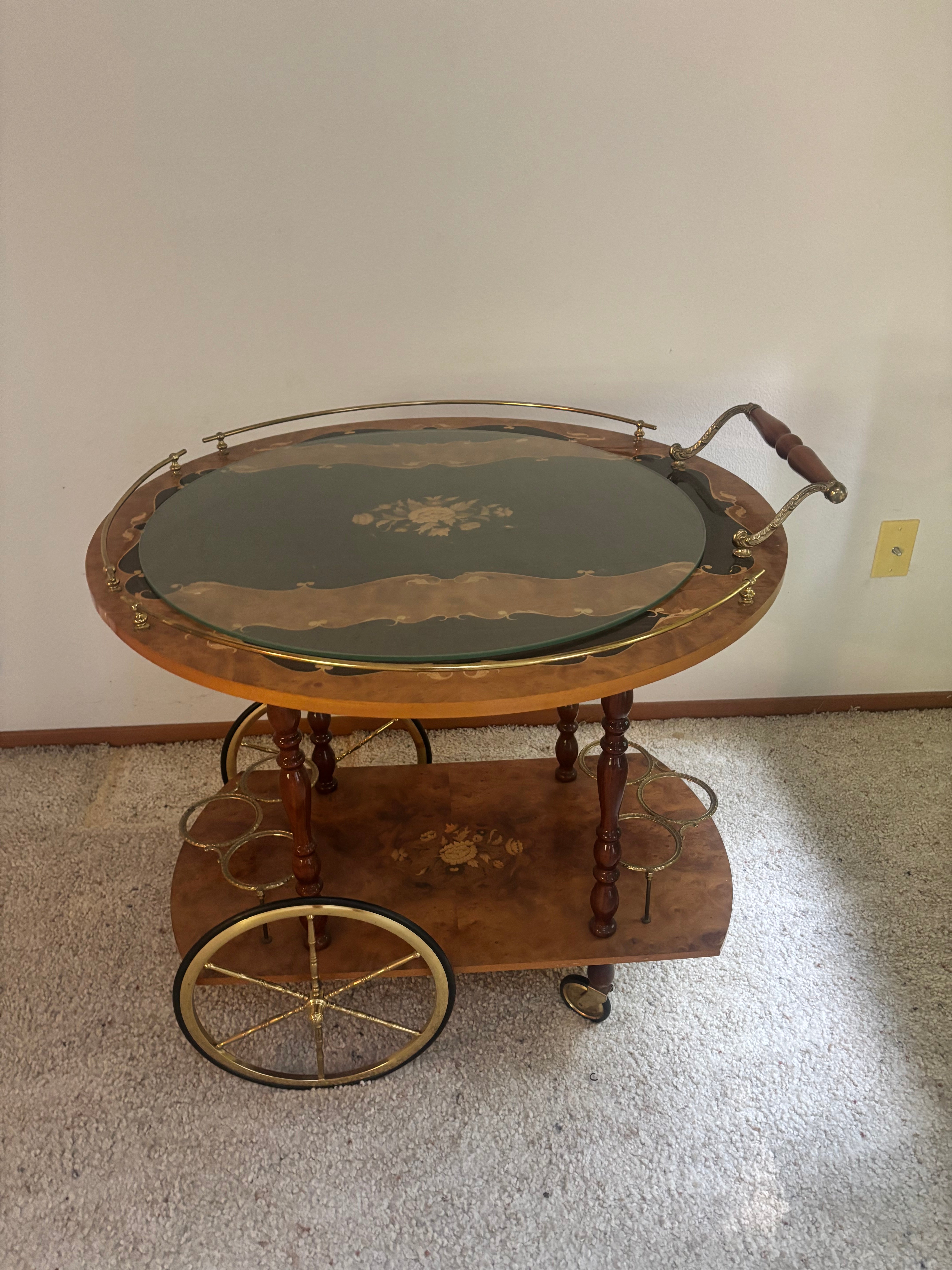 A vintage wooden bar cart with a decorative top, two shelves, and gold accents, featuring round wheels for easy mobility, placed on a carpeted floor.