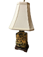 A decorative lamp with a black and gold patterned base and a white fabric shade, resting on a woven surface with a rustic background.
