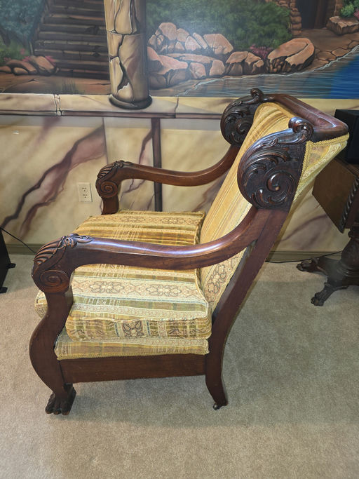 Antique chair with ornate wooden arms and plush yellow patterned upholstery, positioned on beige carpet against a mural backdrop of a natural setting.