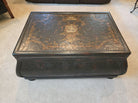 An antique black trunk with intricate artwork on top, featuring ornate patterns and designs. It's placed on a light carpet in a cozy room.