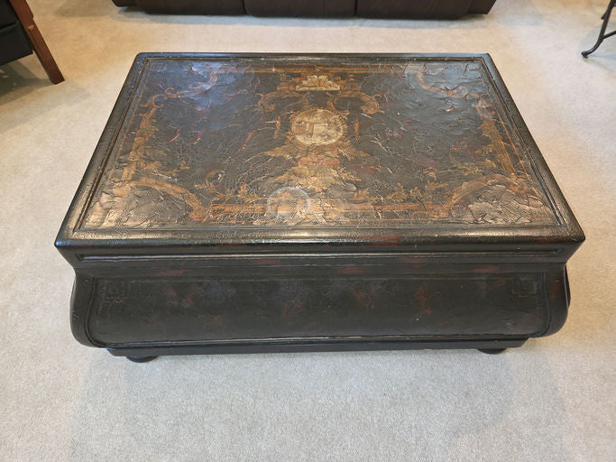 An antique black trunk with intricate artwork on top, featuring ornate patterns and designs. It's placed on a light carpet in a cozy room.