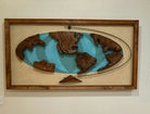 Wooden world map artwork featuring carved continents in earthy tones, set against a turquoise background and framed in wood. 