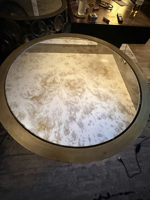 A circular coffee table with a glass top and a patterned, slightly stained surface. Surrounding items include a bottle, glasses, and remote controls.