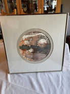 Framed abstract artwork featuring circular designs in earthy tones of orange, gray, and white, displayed on a textured surface.