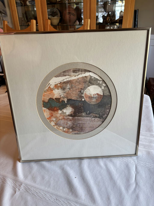 Framed abstract artwork featuring circular designs in earthy tones of orange, gray, and white, displayed on a textured surface.