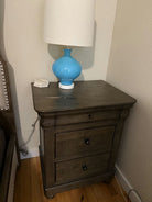 A wooden nightstand with three drawers, topped by a blue lamp with a white shade. A small decorative item sits beside the lamp, on a wooden floor.